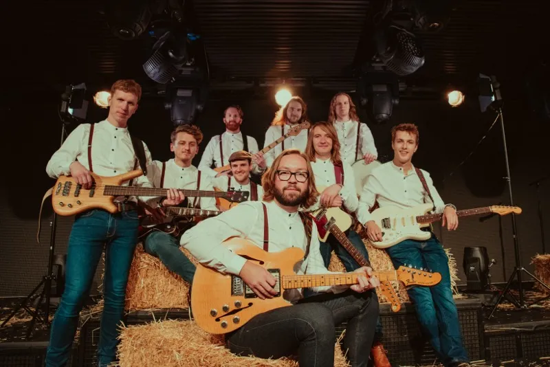 Nine men in white shirts and suspenders posing with electric guitars and basses on hay bales in a dimly lit stage setting.