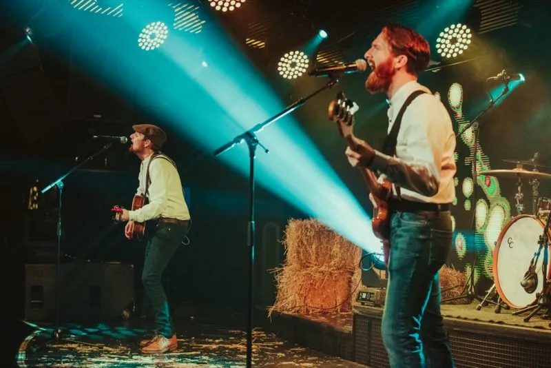 Two musicians performing on stage with guitars, illuminated by blue spotlights and surrounded by hay bales and drums.
