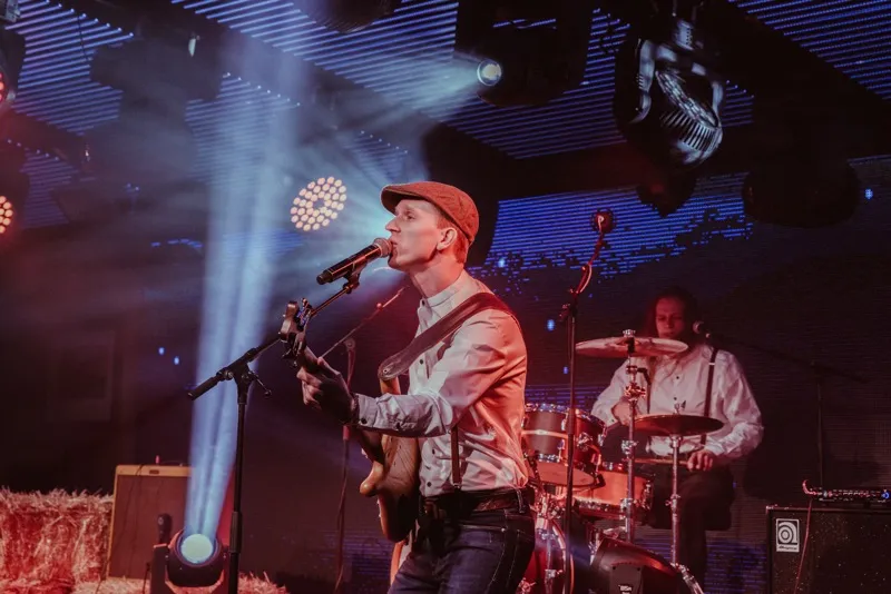 Musician singing into a microphone and playing guitar on stage with a drummer in the background and colorful stage lights.
