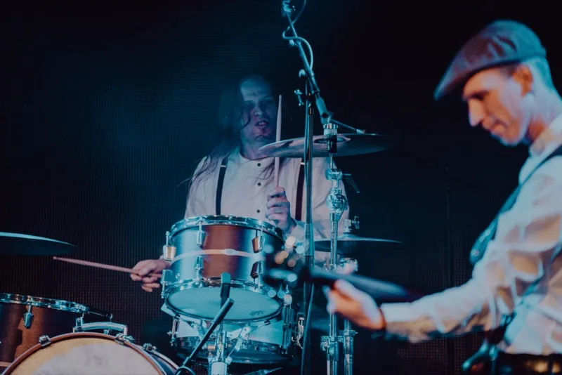Drummer playing a drum set on stage with a guitarist in the foreground wearing a cap.