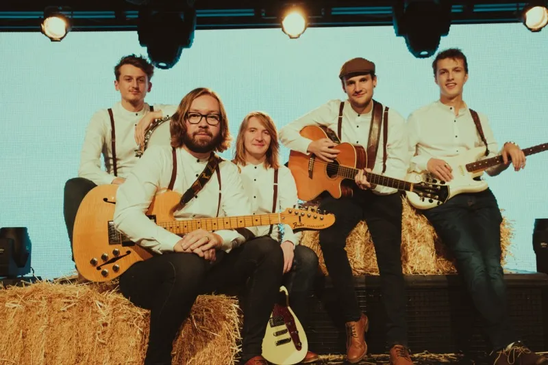 Five young men dressed in white shirts with suspenders posing with guitars on hay bales on a stage.