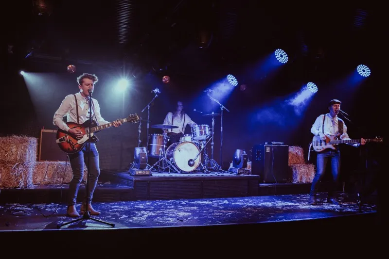 Three-piece band performing on stage with guitars and drums, illuminated by blue stage lights and surrounded by hay bales.
