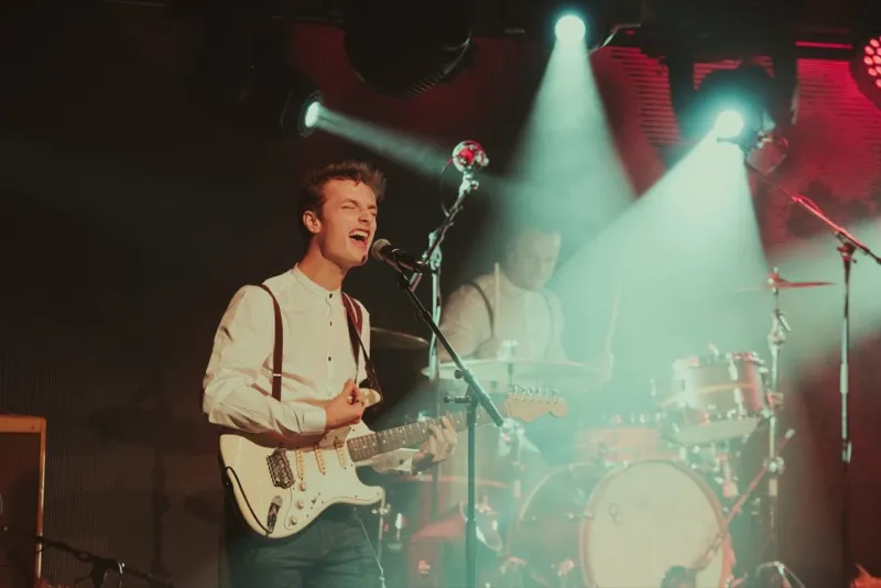 Male guitarist singing into a microphone on stage with drummer playing in the background under bright spotlights.