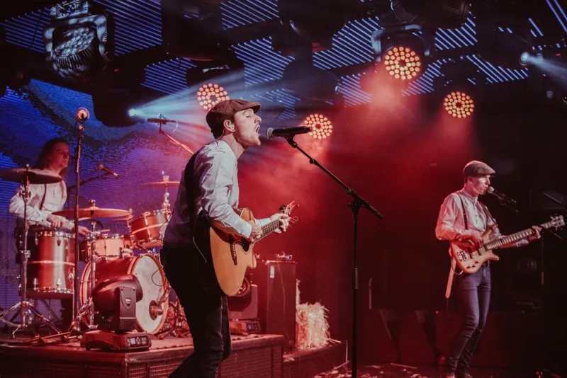 Two musicians playing guitar and singing on stage with a drummer in the background and red stage lighting.