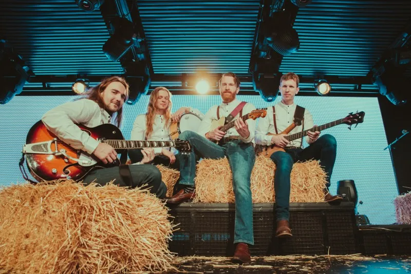 Four male musicians seated on hay bales on a stage, each holding an instrument including guitars and drums, under stage lights.
