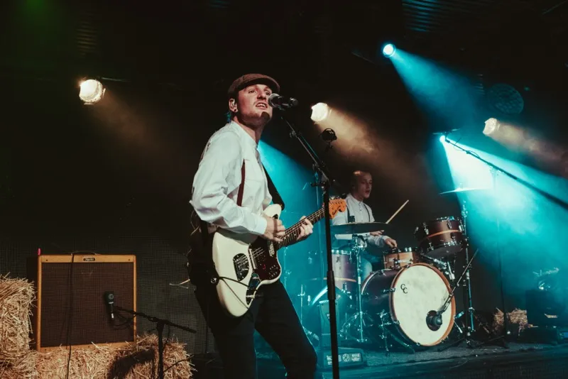 Musician wearing a cap playing electric guitar and singing into microphone with drummer performing under stage lights.