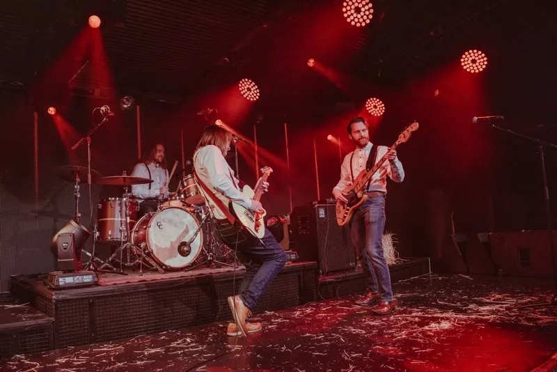 Three musicians performing on stage with red lighting; one playing drums, one playing electric guitar, and one playing bass guitar.