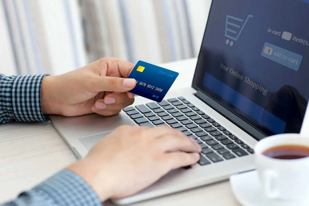 Person holding a blue credit card and using a laptop to shop online, with a cup of tea on the side.