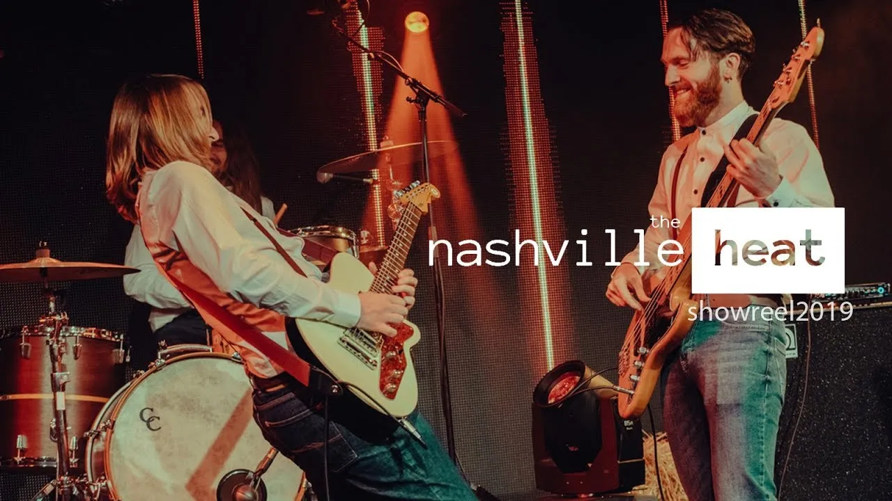 Two musicians playing electric guitar and bass on stage with drums in background under warm orange stage lights.
