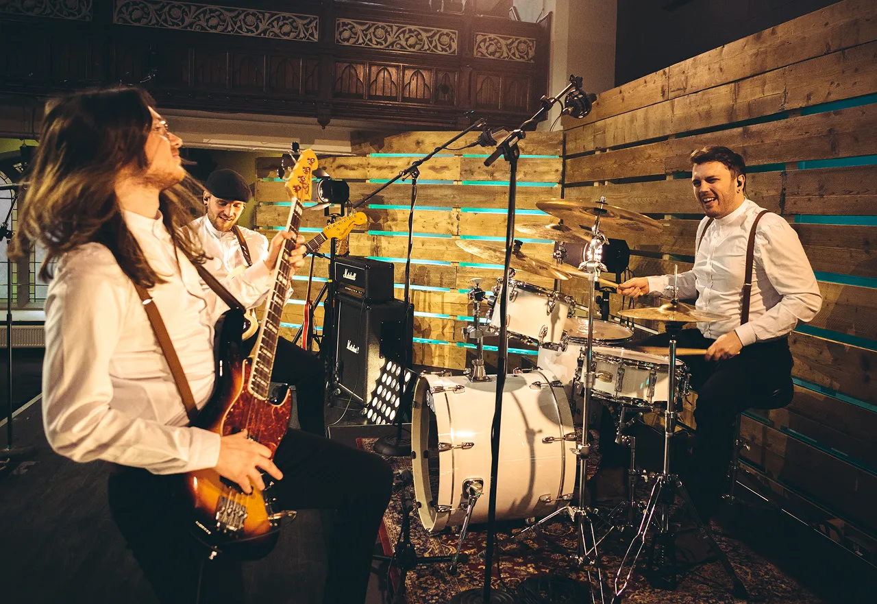 Three musicians performing in a studio with one playing bass guitar, one electric guitar, and one on drums.