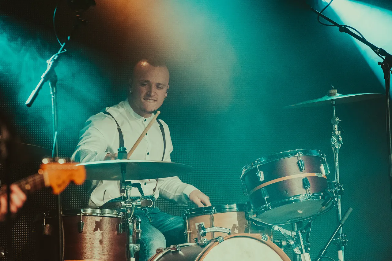 A drummer in a white shirt and suspenders playing drums on stage under blue and orange lighting.
