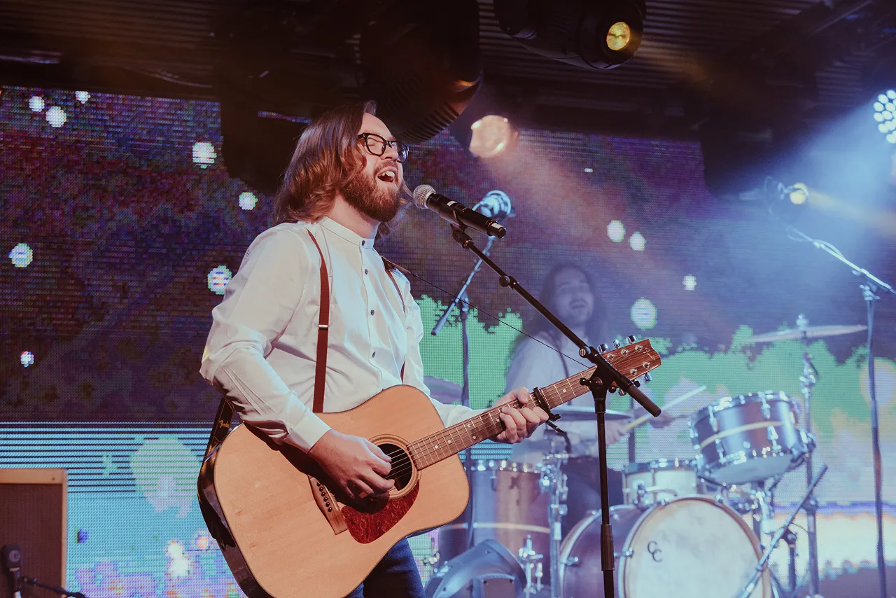 Bearded male musician with glasses singing and playing acoustic guitar on stage with drummer in the background.