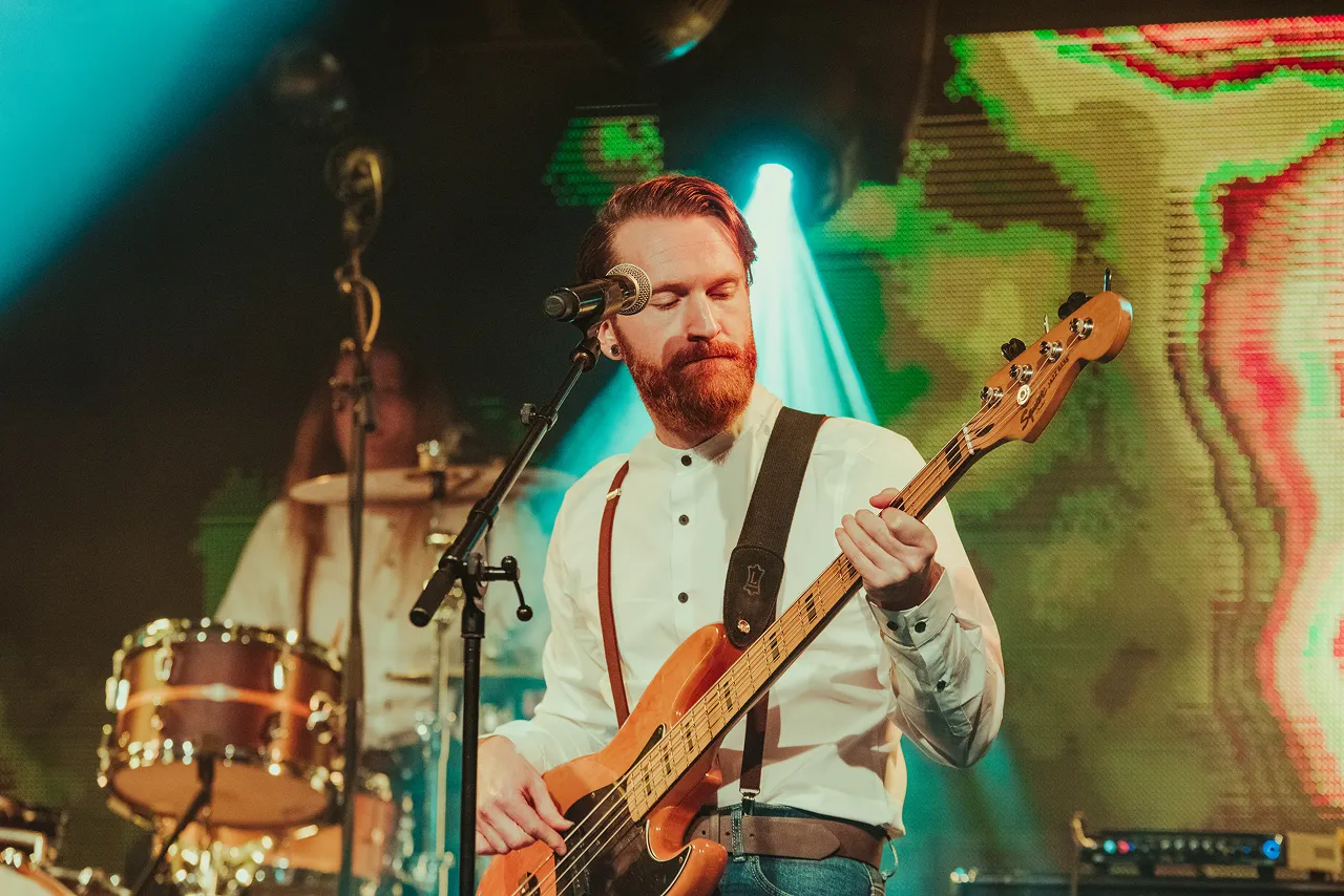 Bearded musician playing bass guitar on stage with microphone and drummer in background under blue stage light.
